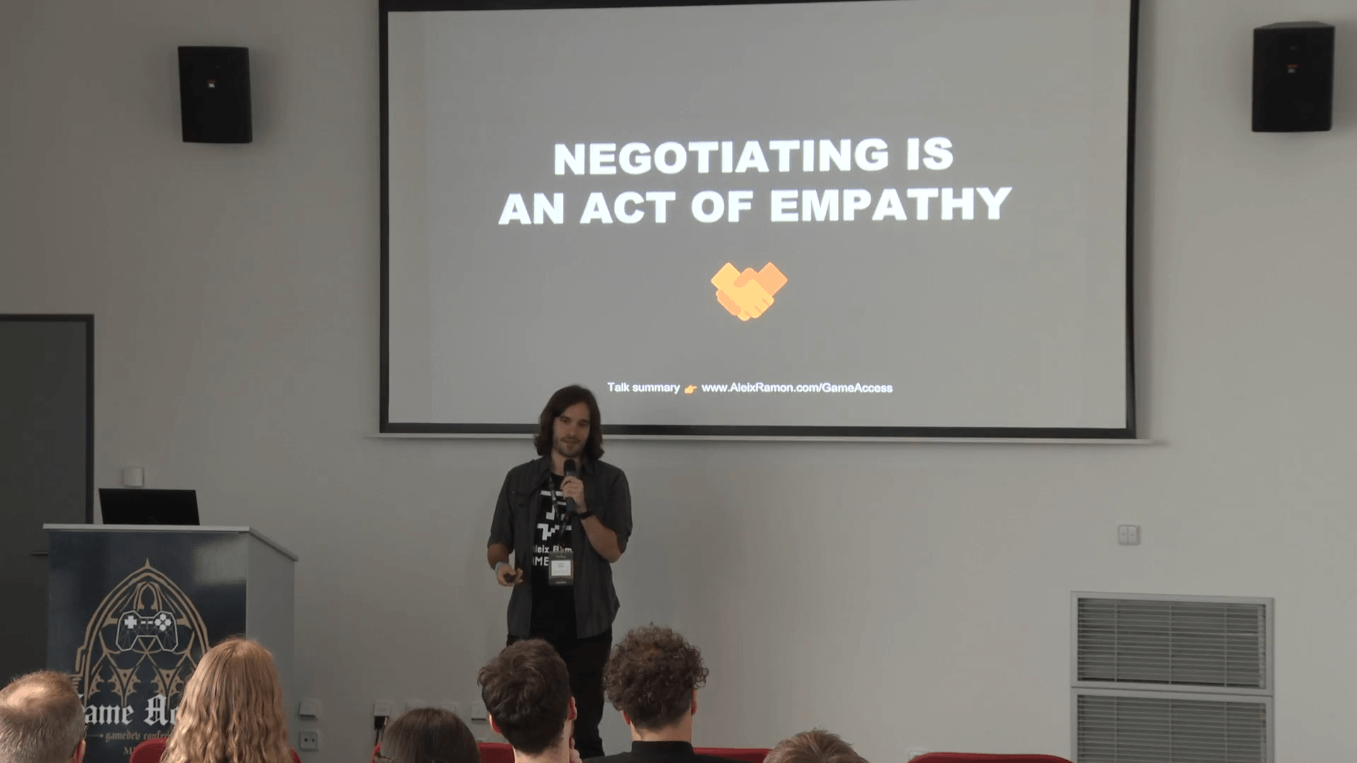 Speaker presenting a talk with slide titled 'Negotiating is an act of empathy' and a handshake icon in a conference room.