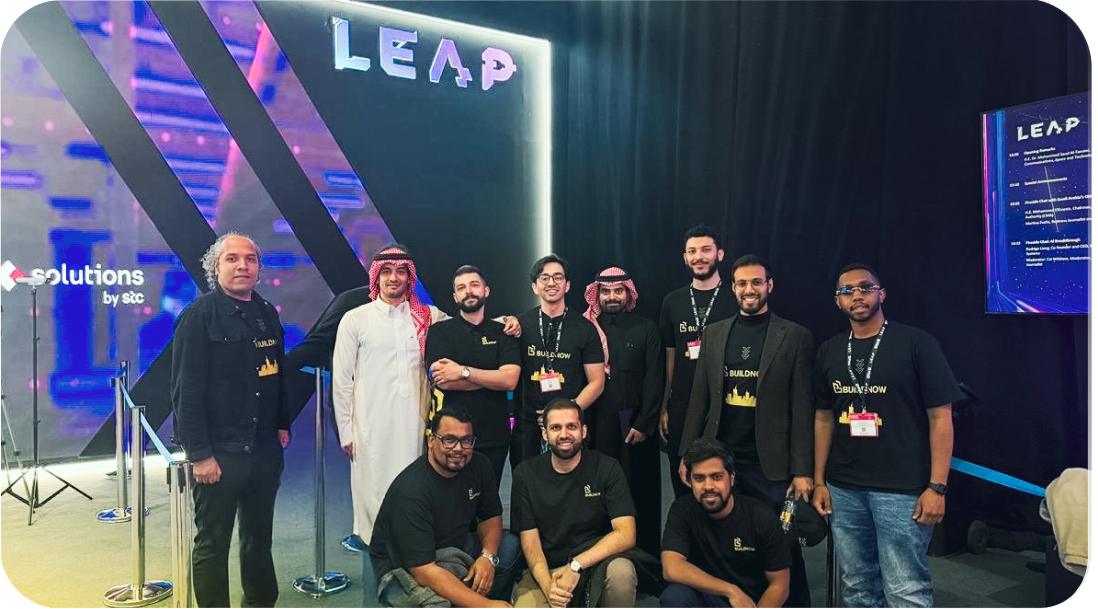 مجموعة من الرجال يقفون يبتسمون في حدث LEAP مع شعار BuildNow وشعار solutions by stc في الخلفية.