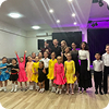 Group of children and adults posing together in colorful dance costumes on a wooden floor under stage lighting.