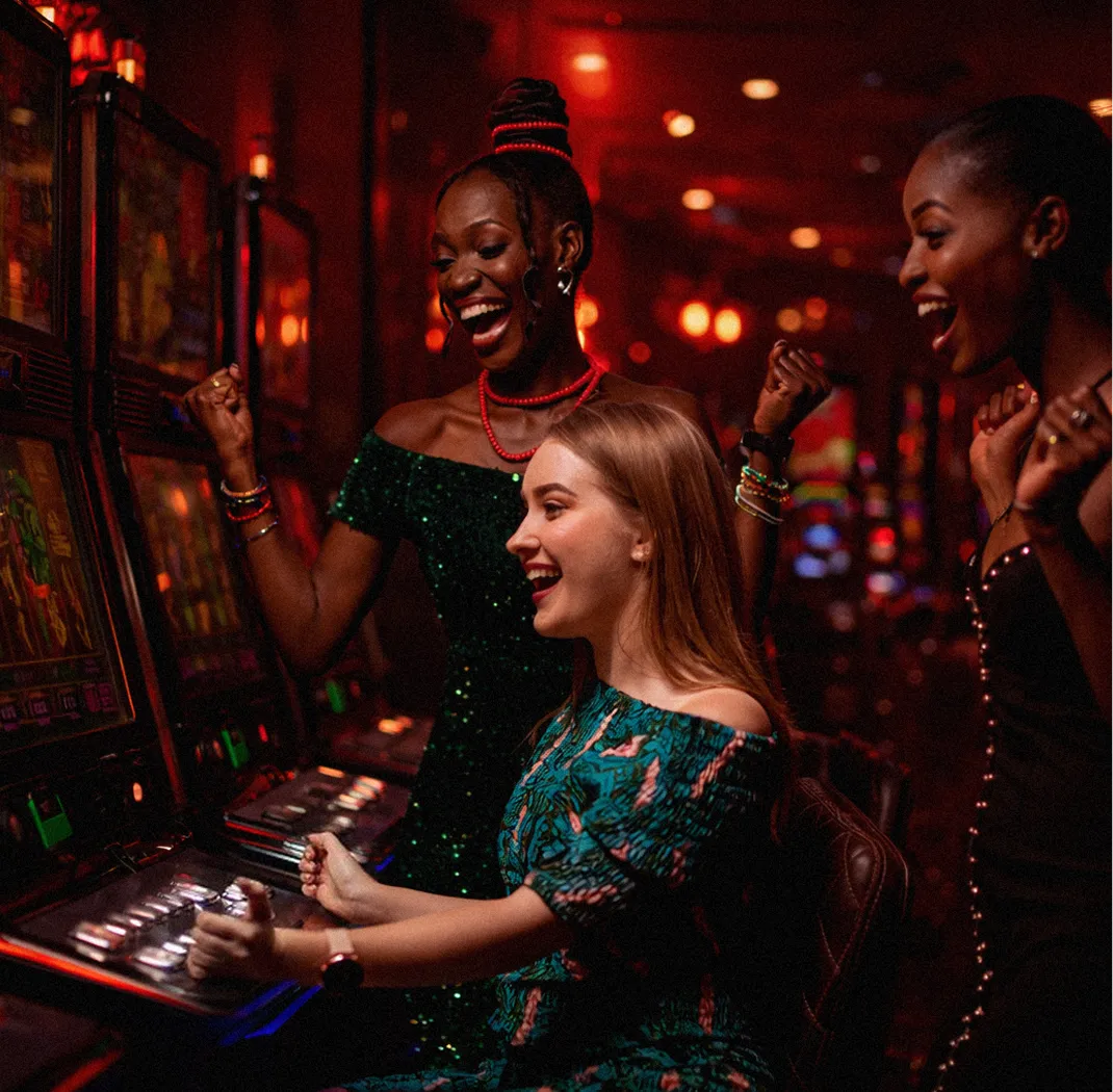 Three women celebrating a win at a casino slot machine, smiling and cheering in low red lighting.