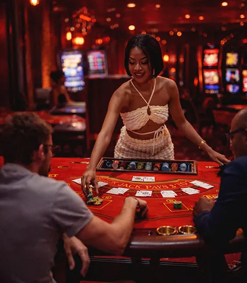 A female casino dealer in a beige outfit managing chips at a blackjack table with two male players seated.