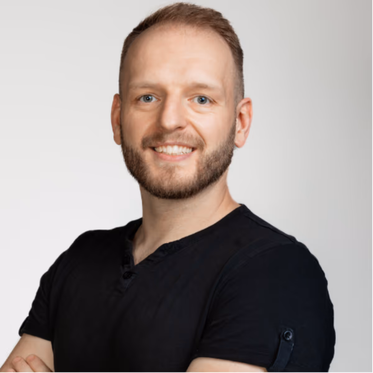 Uśmiechający się mężczyzna z brodą i krótkimi blond włosami w czarnej koszulce, stojący na jasnoszarym tle.