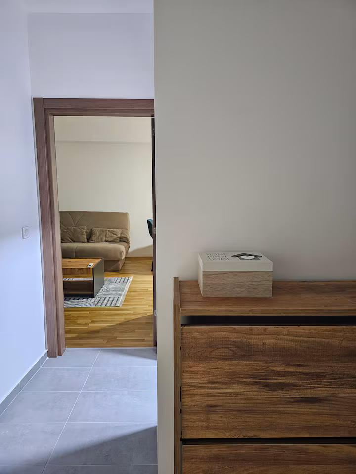 Interior view of a hallway leading to a living room with a beige sofa and a wooden coffee table on a patterned rug, and a wooden cabinet with a box on it in the foreground.