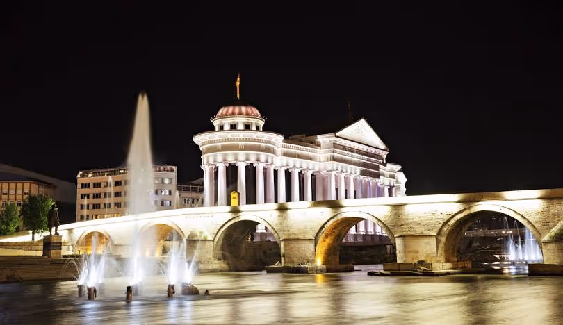 Night view of a historic bridge with arches and fountains in the water, illuminated classical building in the background.