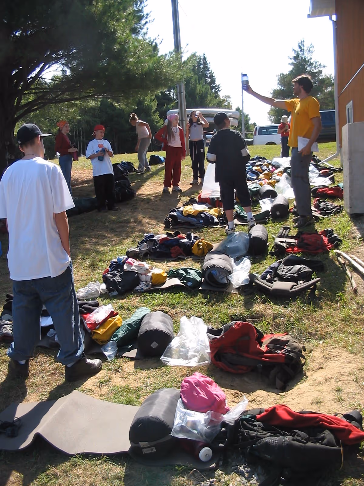 Group of people outdoors preparing camping gear including rolled sleeping bags, backpacks, and clothing spread out on grass.