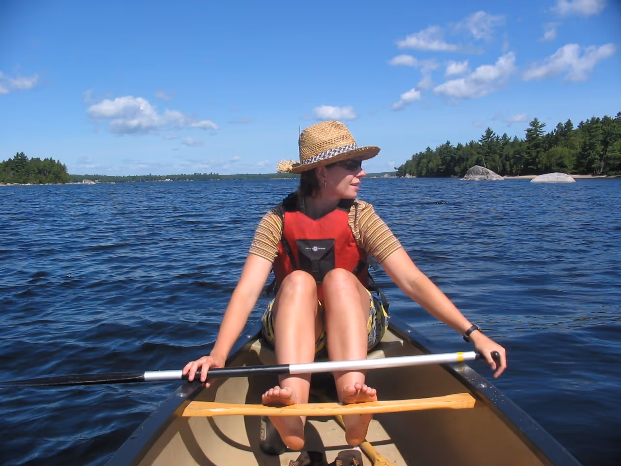 Woman wearing a straw hat and red life vest sitting barefoot in a canoe on a lake with trees and rocks in the background under a blue sky.