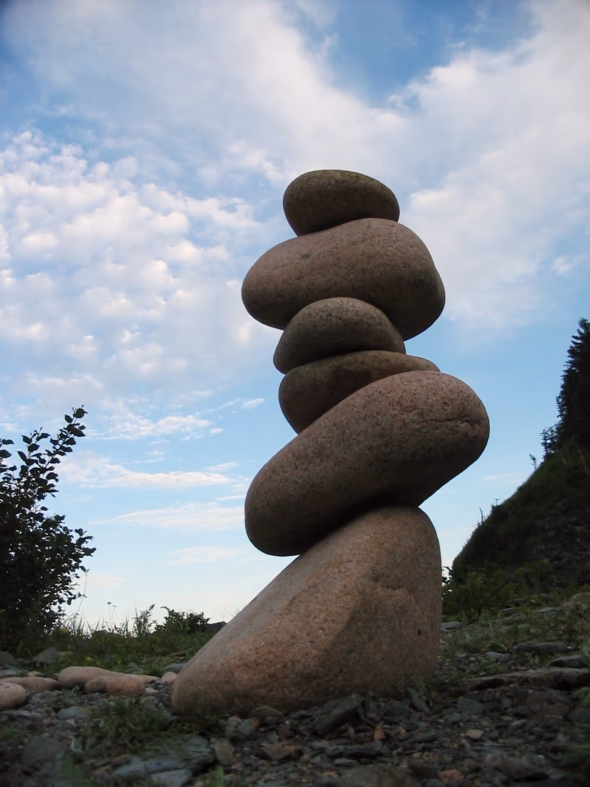 Stack of six smooth rounded stones balanced on rocky ground under a partly cloudy sky.