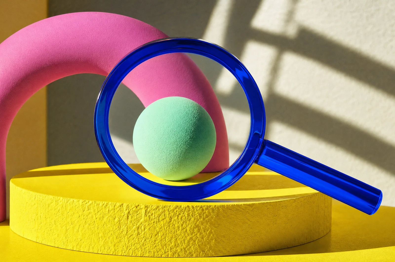 A textured green sphere centered on a yellow circular platform with a pink arch behind it, viewed through a large blue magnifying glass casting shadows on a cream wall.