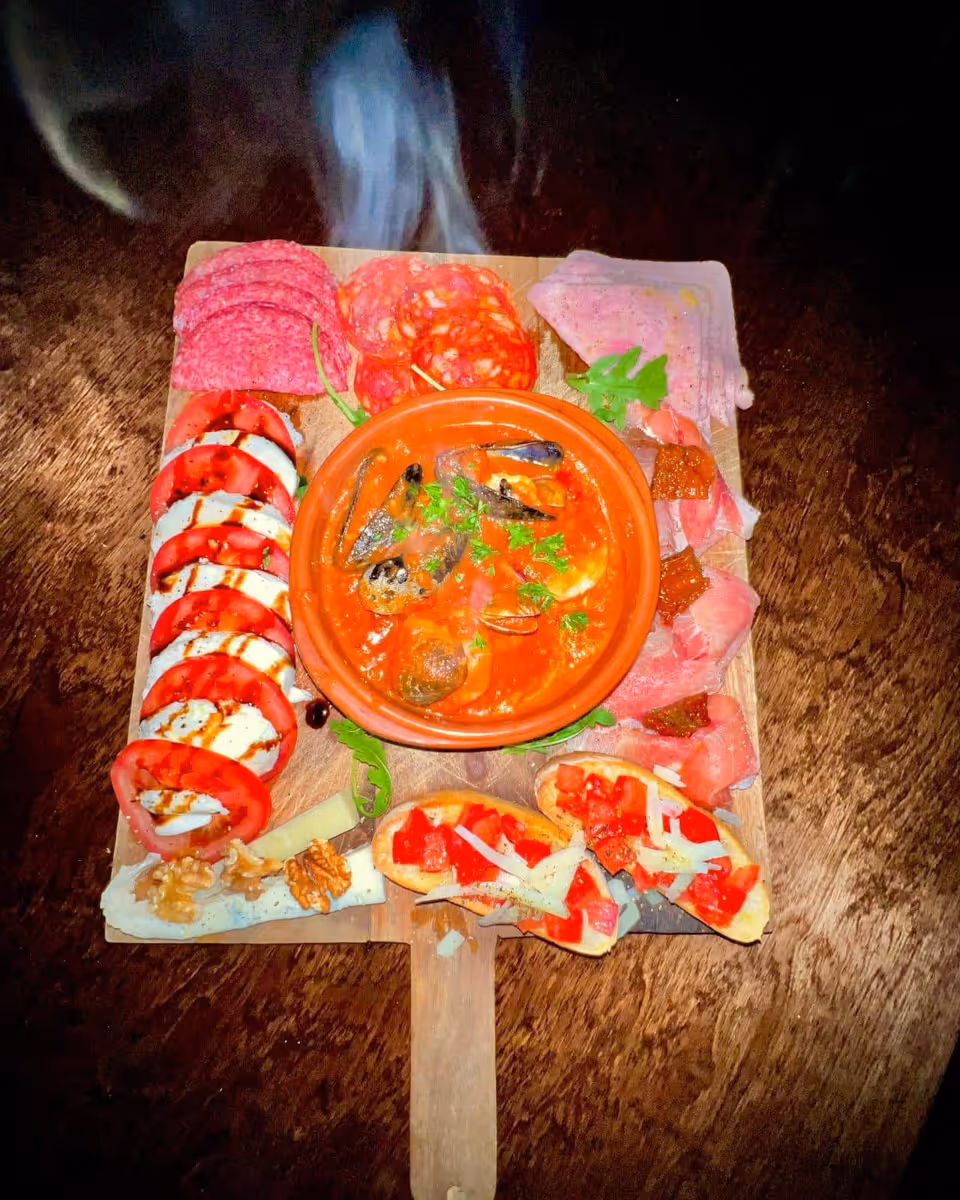 Houten tapasplank met schotel dampende mosselen in tomatensaus, verschillende fijne vleeswaren, plakjes tomaat en mozzarella met balsamico, kaas met walnoten, en bruschetta met tomaat en kaas.