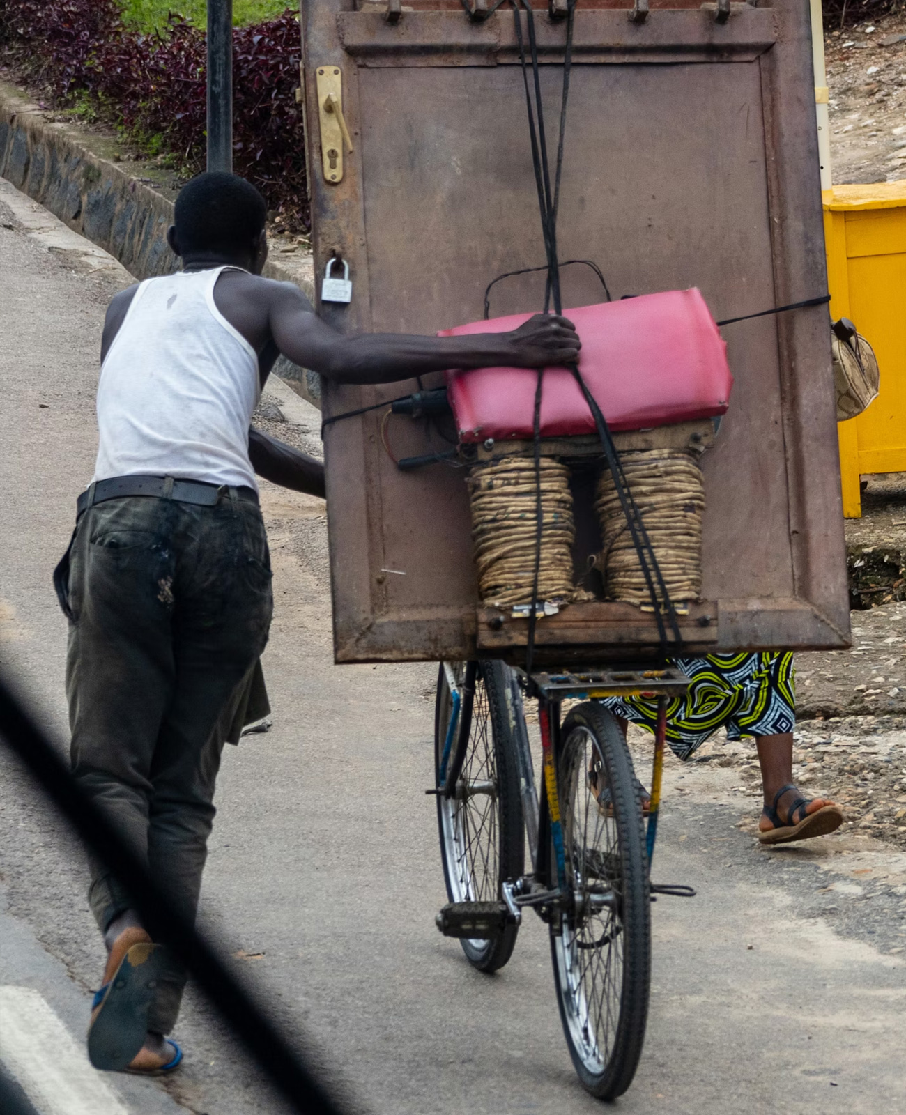 Man pushing a bicycle loaded with a large wooden board and baskets on a paved road.