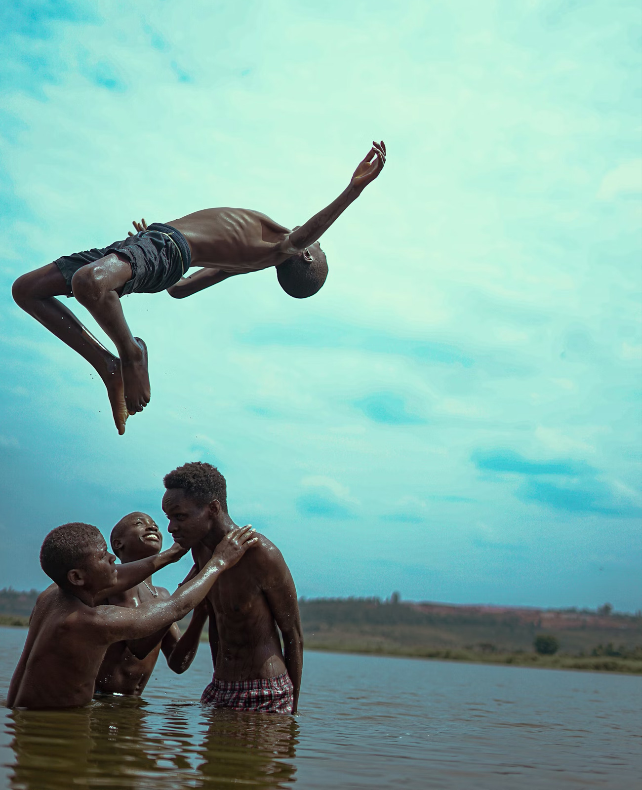 Four boys playing in a lake, with one boy flipping backward in mid-air above three others standing in the water.