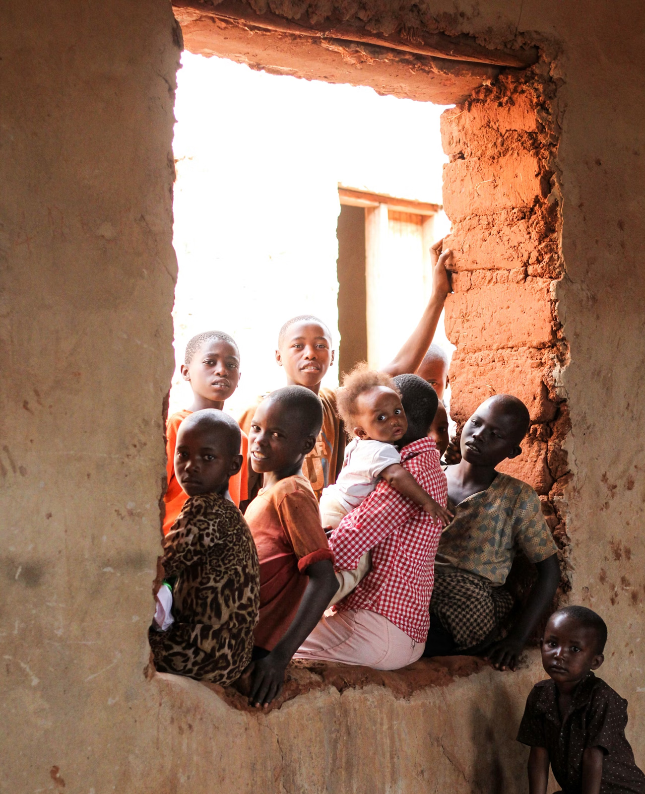Group of children sitting and standing in a rustic window frame of a mud brick building, one child holding a baby.