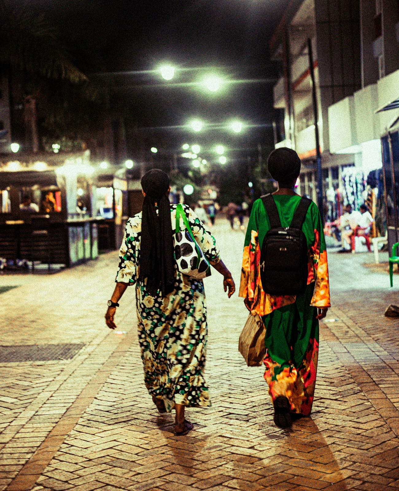 Two women wearing colorful dresses walking on a lit cobblestone street at night.