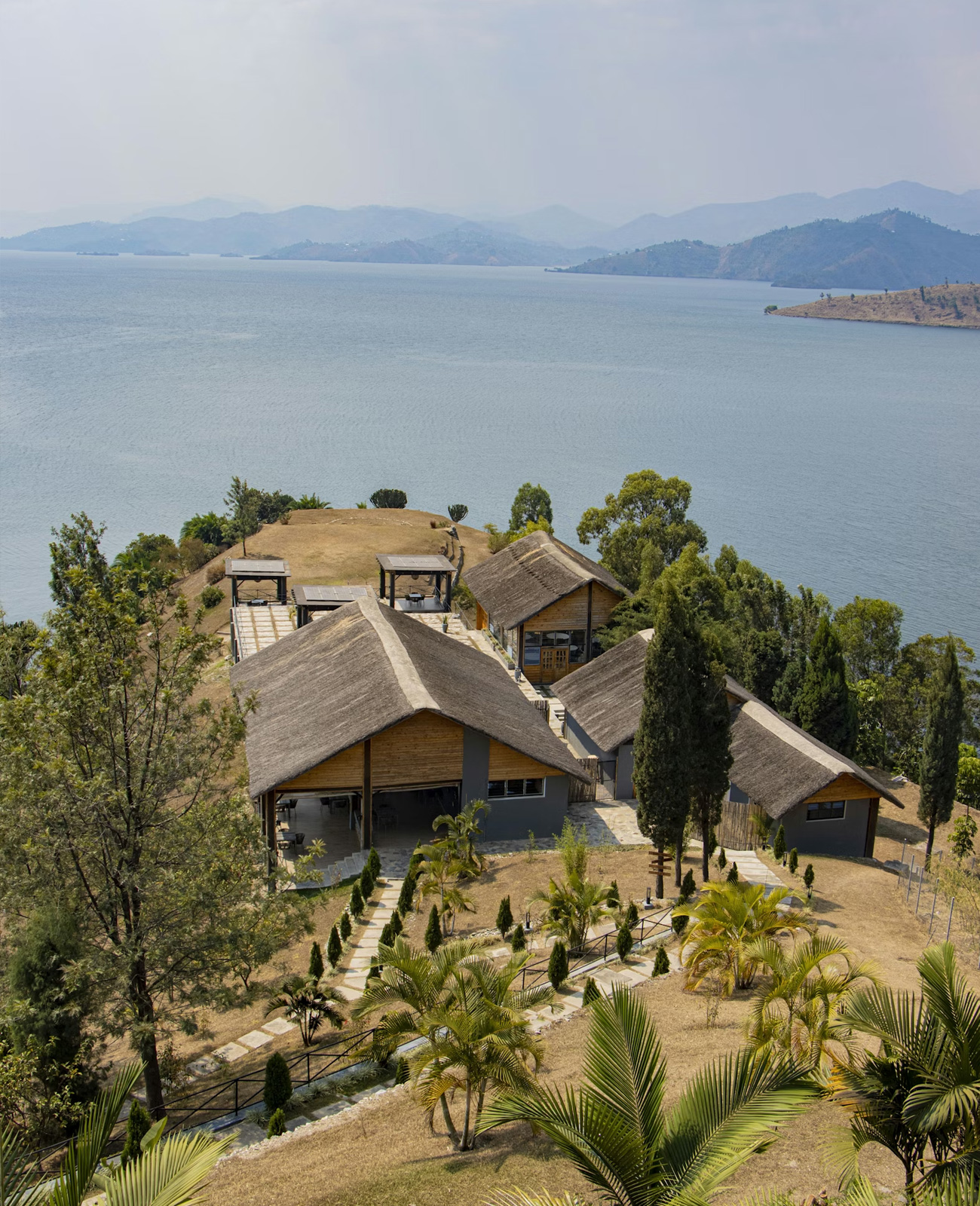 A lakeside resort with thatched roof buildings surrounded by trees and pathways, overlooking a large lake with distant hills.