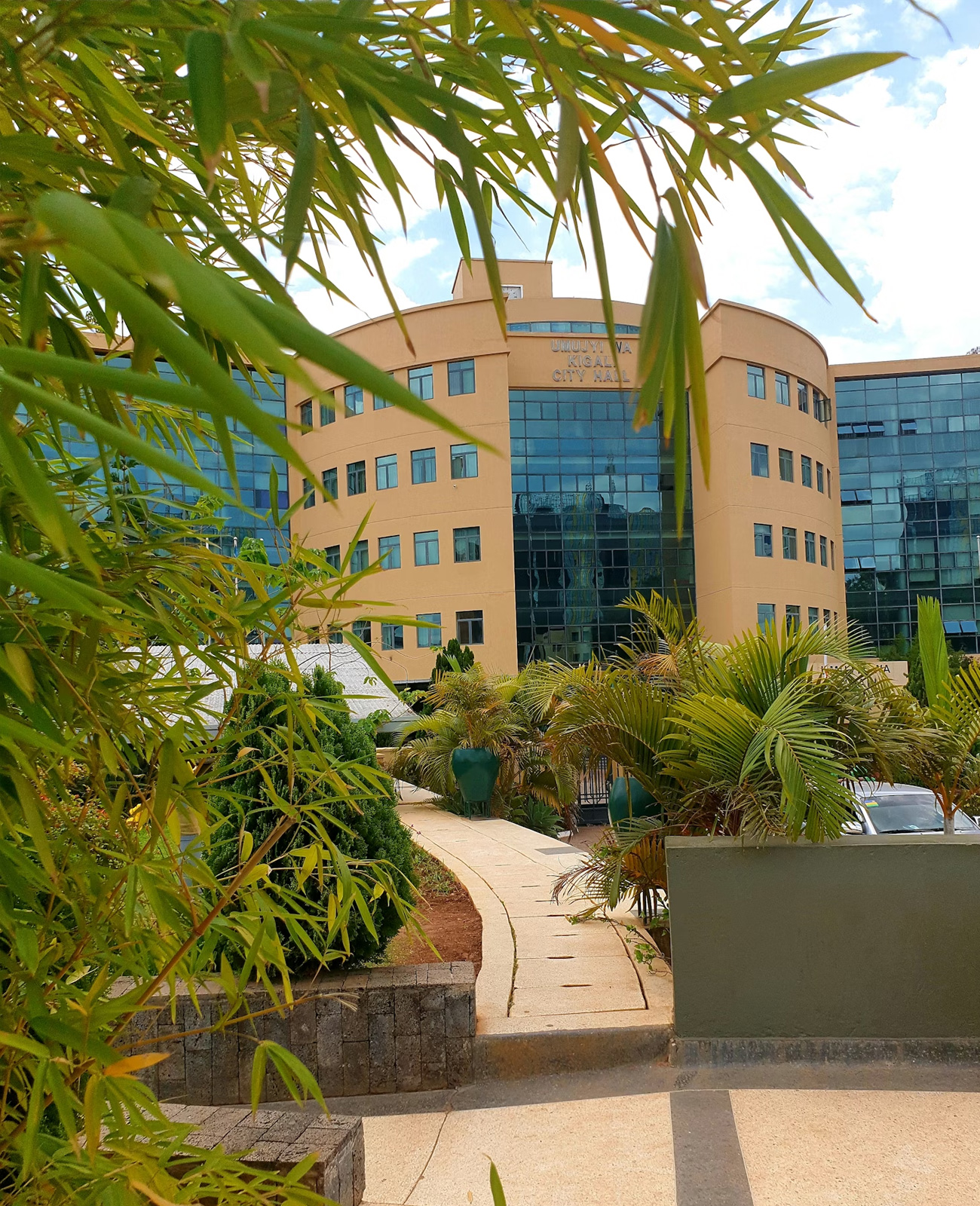 Pathway lined with green plants leading to Kigali City Hall, a modern building with curved beige walls and large blue-tinted windows.