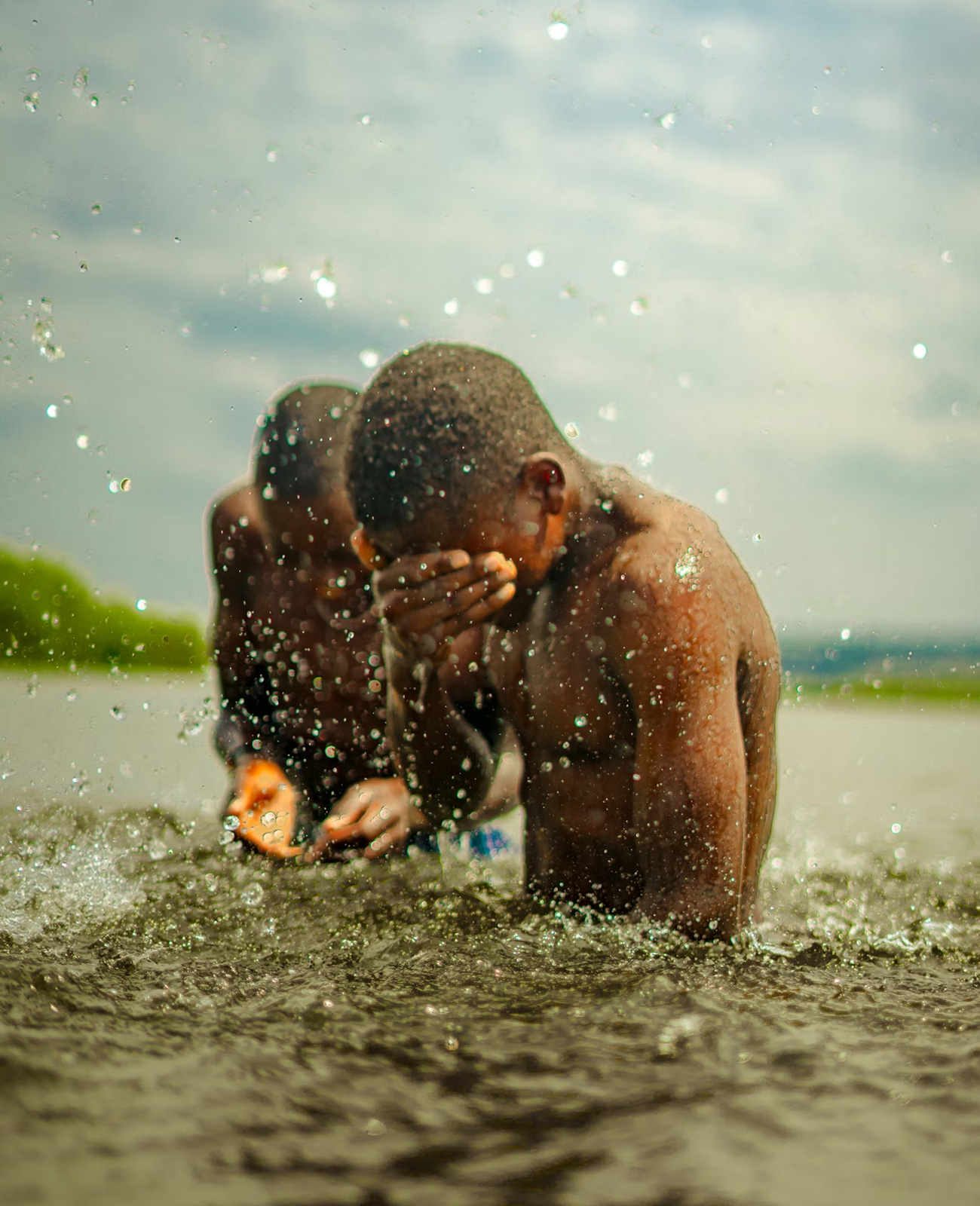 Two boys splashing and playing in shallow water outdoors under a cloudy sky.