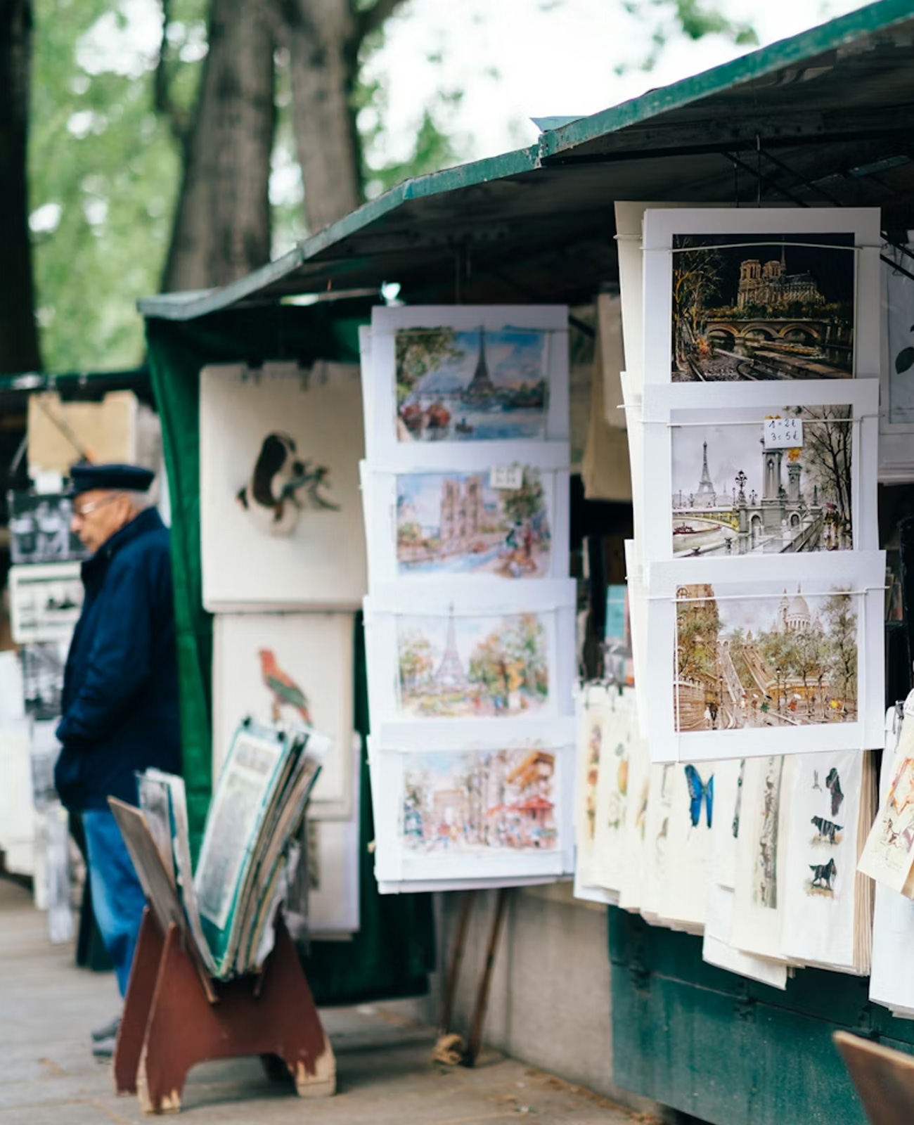 Outdoor art stall displaying various paintings and sketches of Paris landmarks with a man in a blue jacket and hat standing nearby.