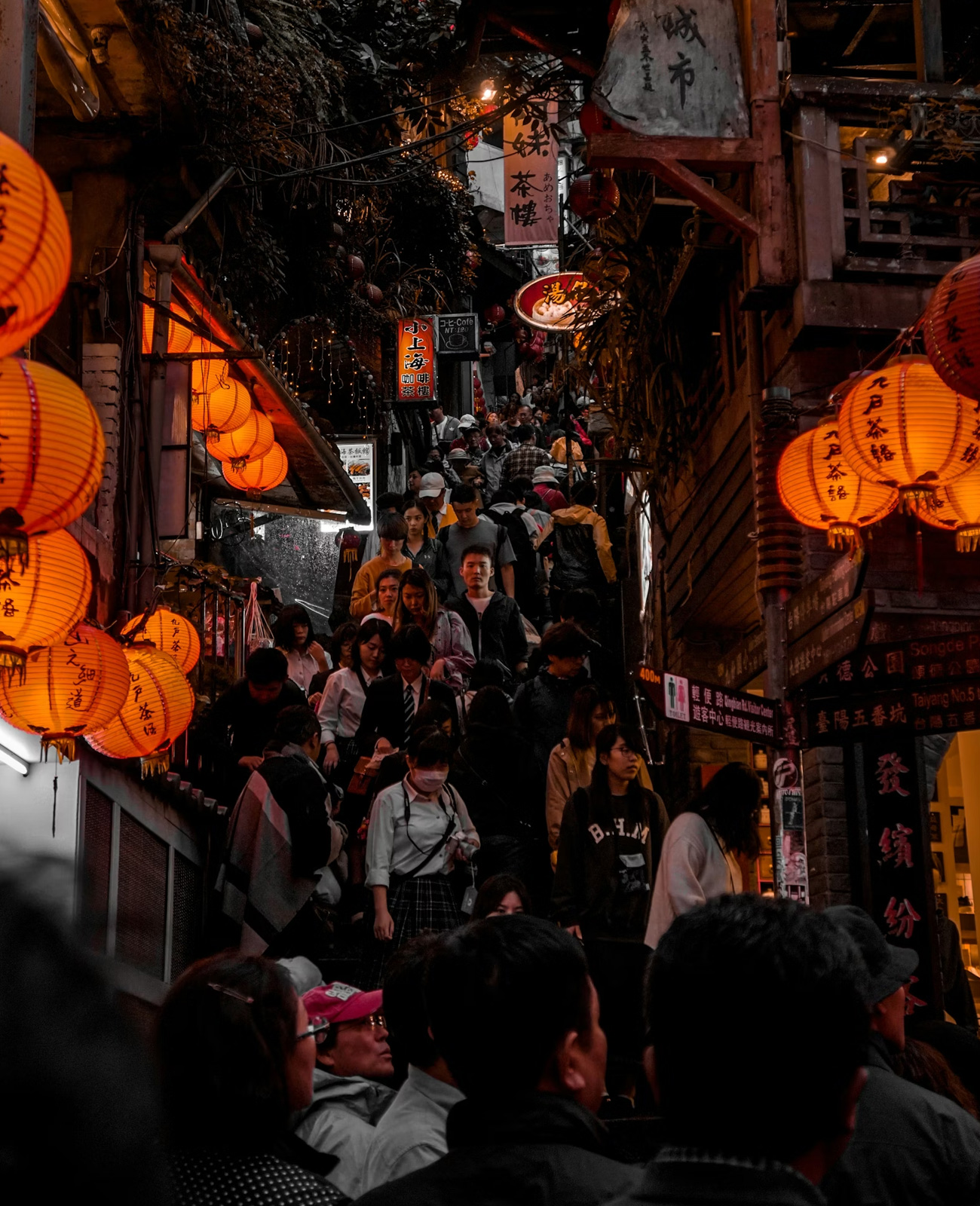 Crowded narrow street in an Asian city at night with glowing orange lanterns and many people walking on stairs.