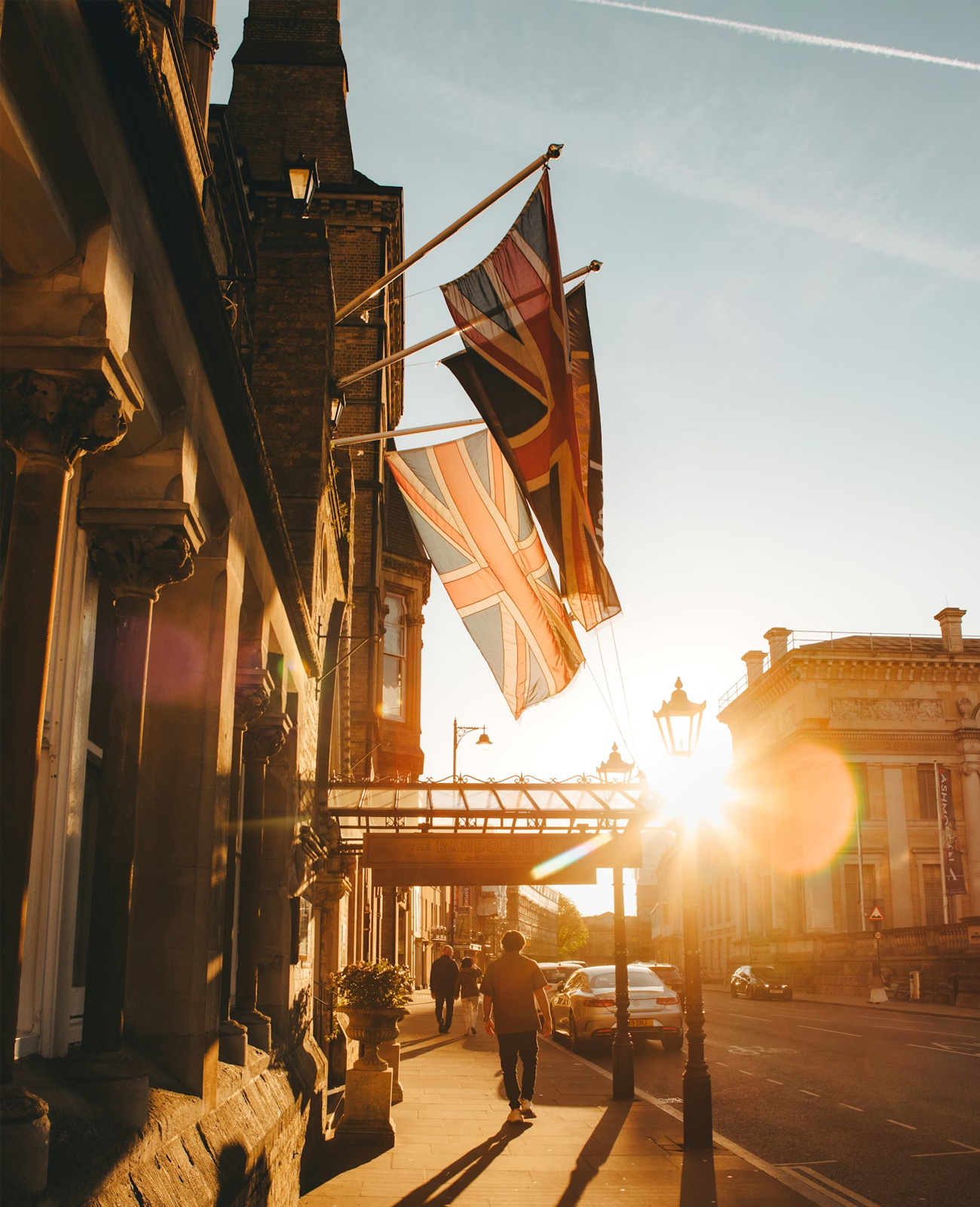 Sunlit street scene with Union Jack flags hanging from a building and pedestrians walking on the sidewalk.