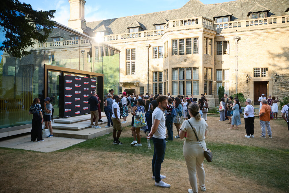 Historic stone building with gothic windows and spires, two people walking along a garden path beside green lawn.