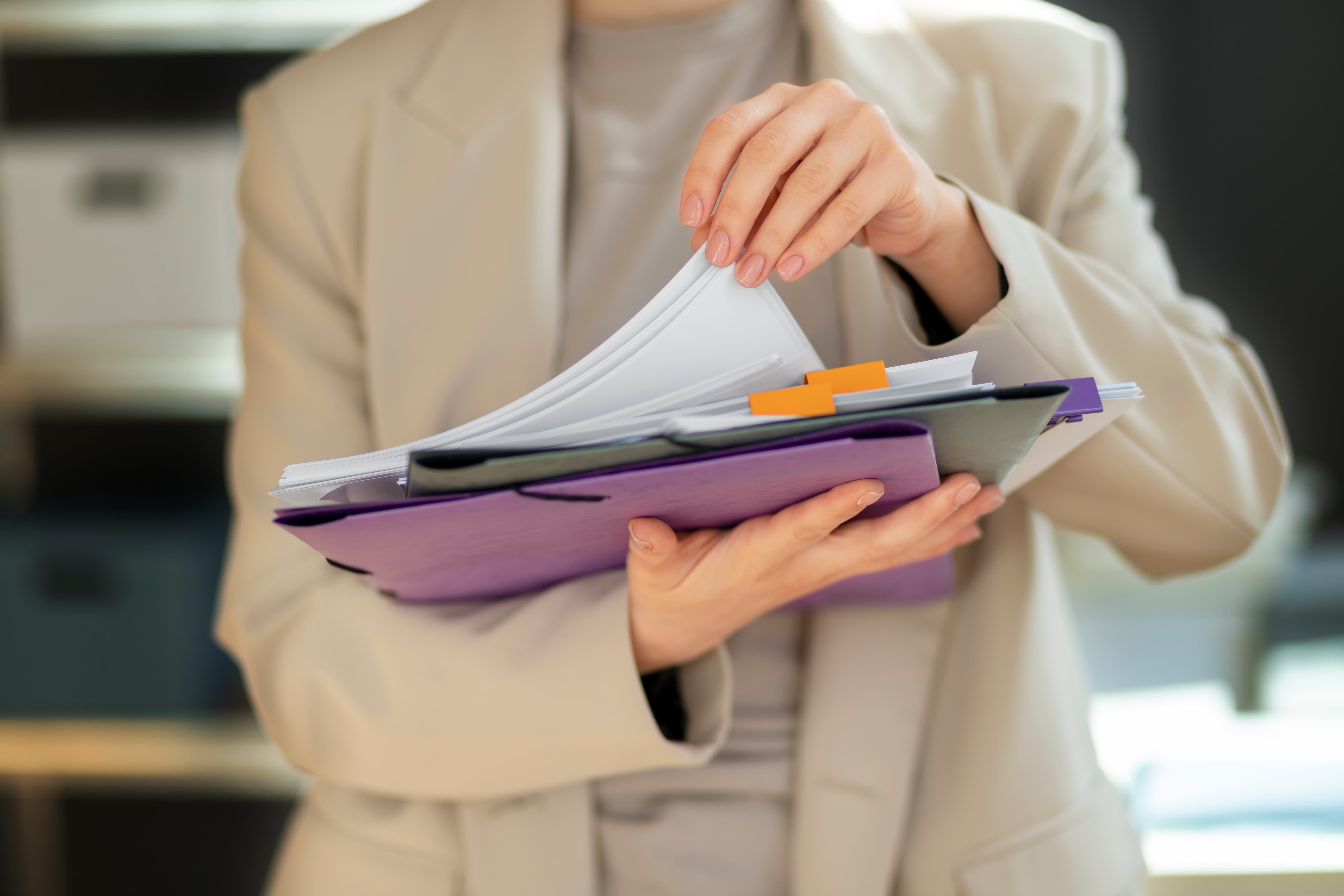 Person in beige blazer organizing papers and folders with colored tabs.