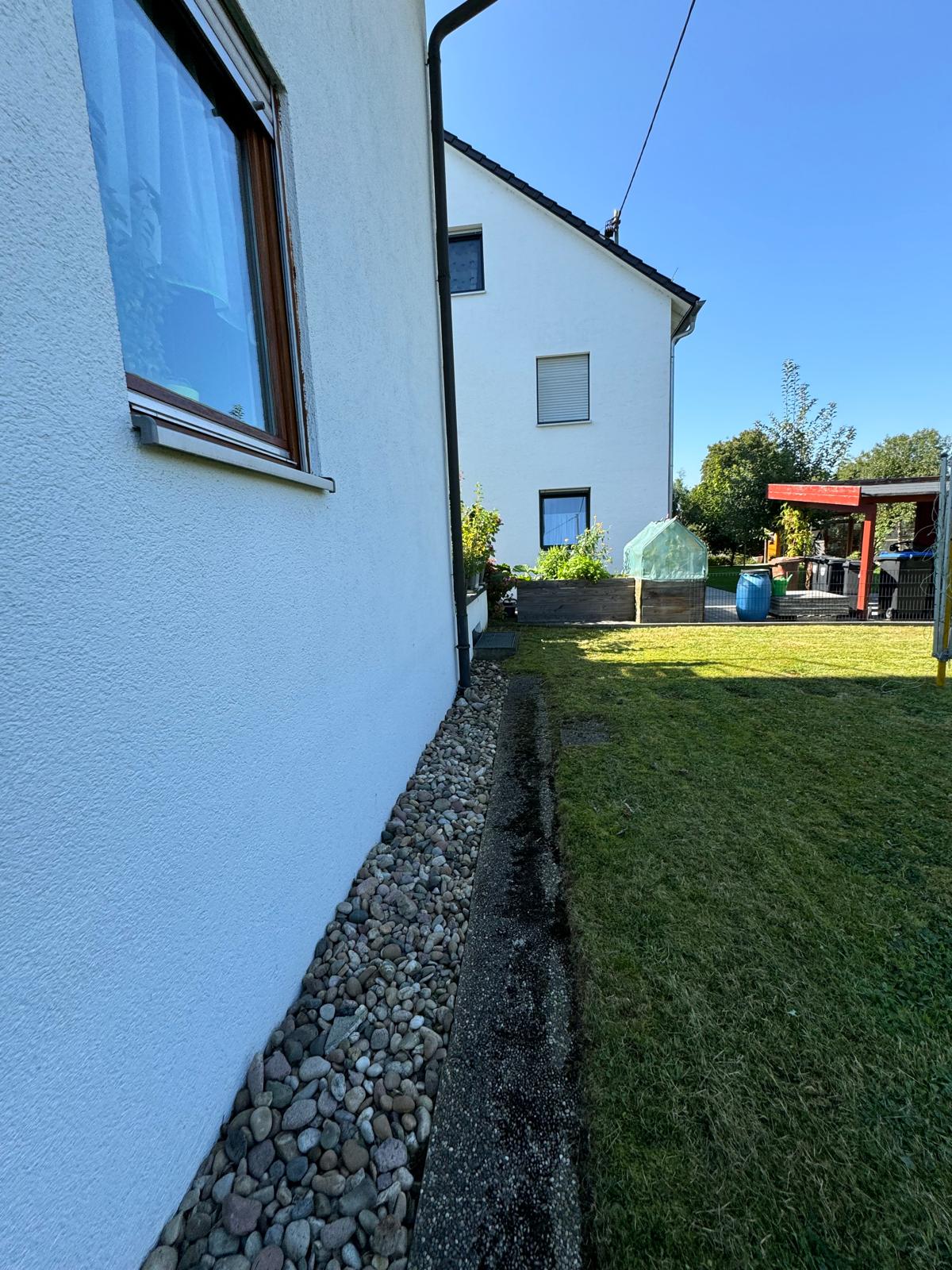 Gardener wearing beige overalls and green cap kneeling on grass while trimming a shrub with hedge clippers in a lush garden.