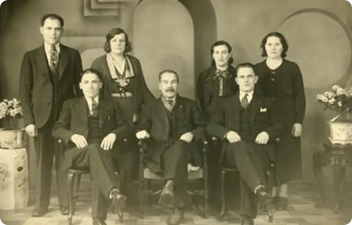 Black and white vintage group portrait of three seated men and four standing women and men, dressed formally in suits and dresses.