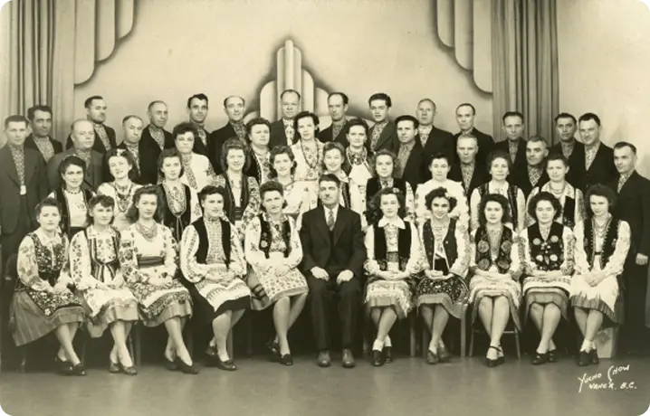 Black and white group photo of men and women, with women mostly in traditional embroidered dresses and men in suits, posed indoors against an art deco style backdrop.