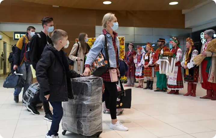 People wearing face masks with luggage walking through an airport terminal where a group in traditional Eastern European attire welcomes them.