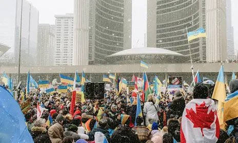 Large crowd holding Ukrainian flags and signs in a snowy urban setting with tall buildings.
