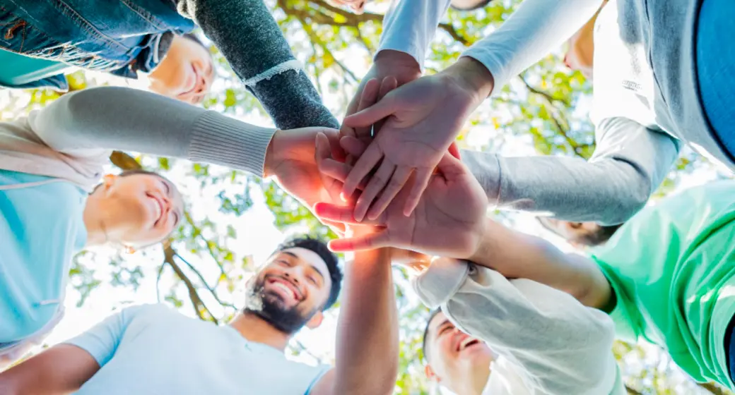 Group of diverse volunteers smiling and stacking their hands together outdoors under trees.