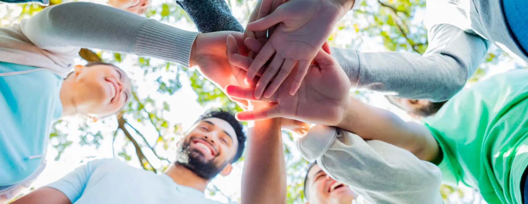 Group of diverse young people standing in a circle outdoors with their hands stacked together in the center.
