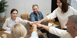 Five colleagues in an office putting their hands together in a teamwork gesture around a table.