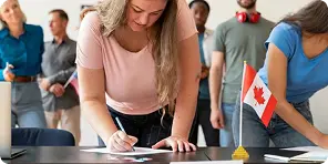 Young woman with long hair signing a document on a table with a small Canadian flag, while people wait in line behind her.