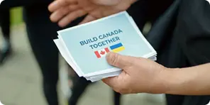 A person holding flyers that read 'BUILD CANADA TOGETHER' with Canadian and Ukrainian flags.