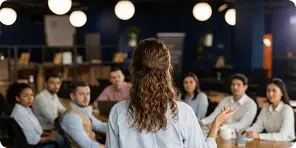 Woman speaking and gesturing to a diverse group seated at a long table in a modern meeting room.