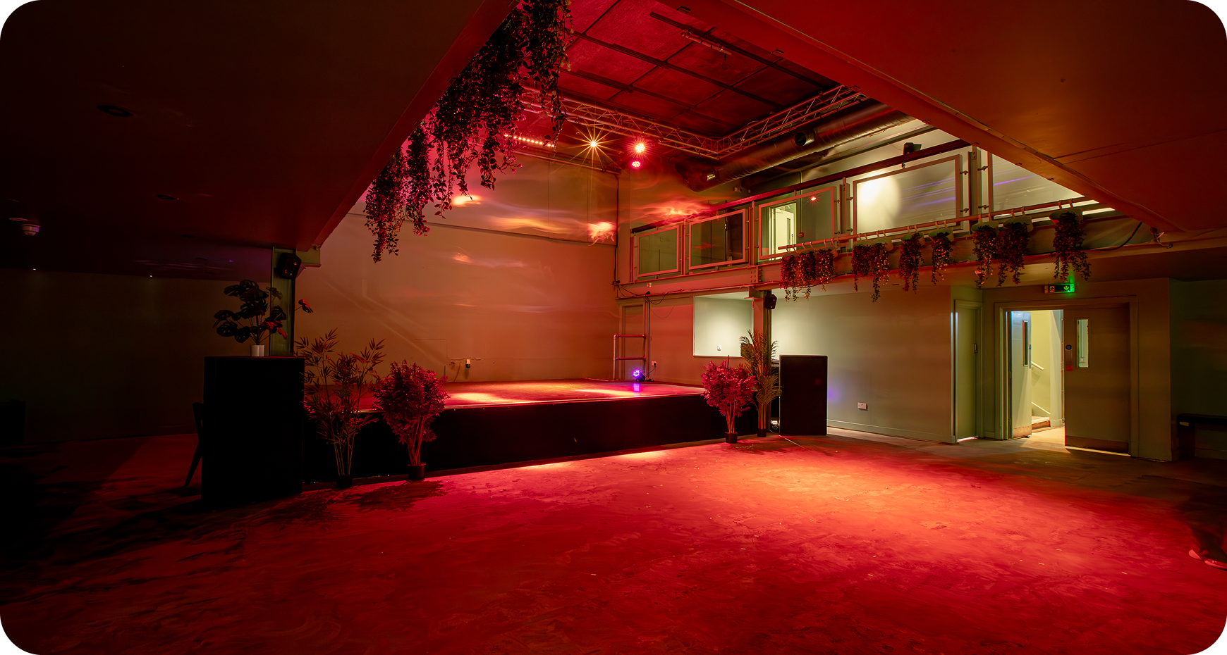 Empty indoor event space with a stage lit by red and orange lights, decorated with hanging and potted plants.