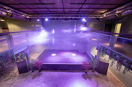 Empty stage in an indoor event space with purple lighting and fog effects, surrounded by railings and decorated with plants.