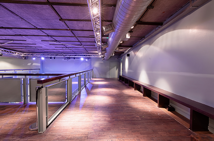 Empty indoor corridor with wooden floor, metal railing, bench seating along the wall, and exposed industrial ceiling with ventilation ducts.