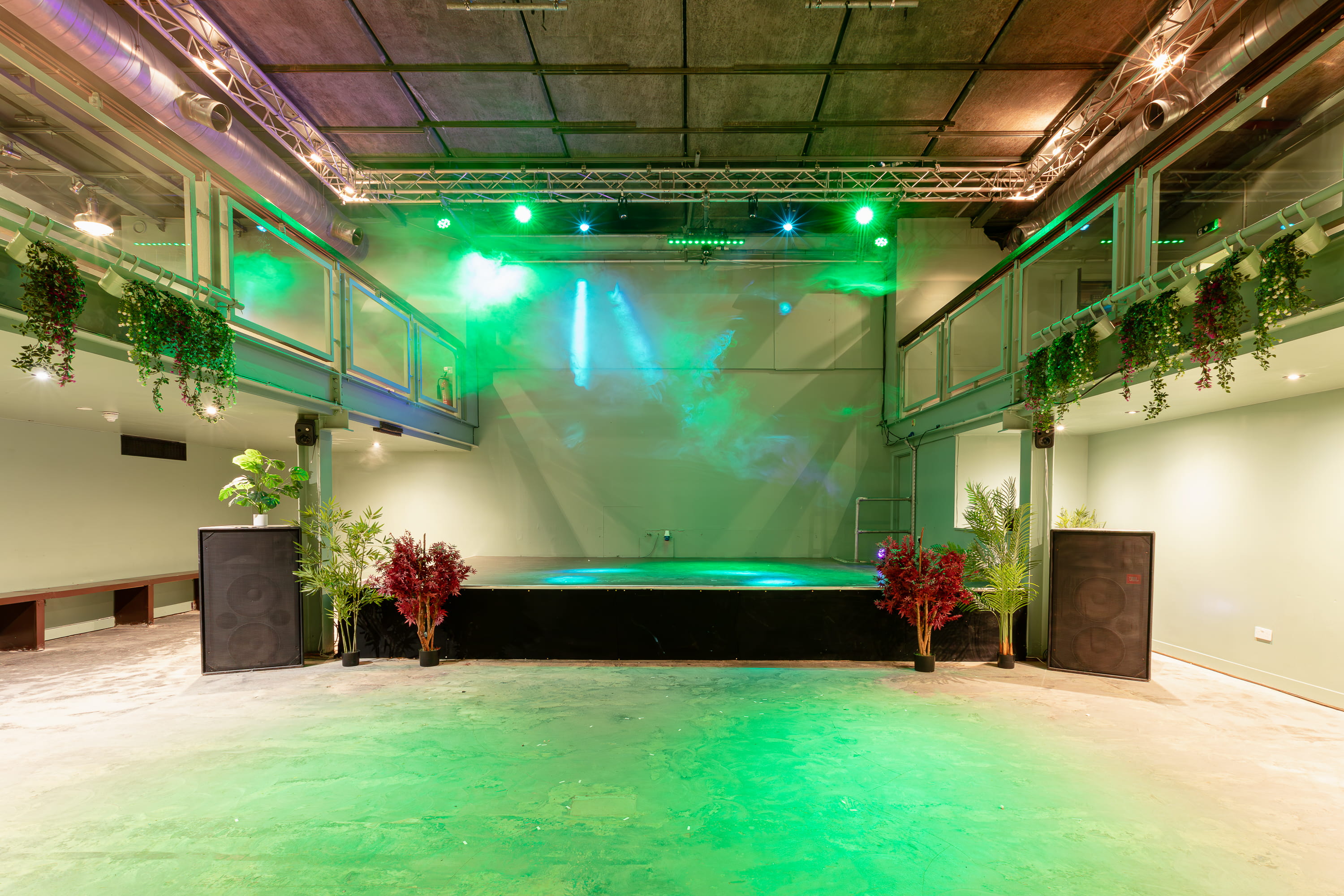 Empty indoor stage with green and blue stage lights, surrounded by potted plants and large speakers on a concrete floor.