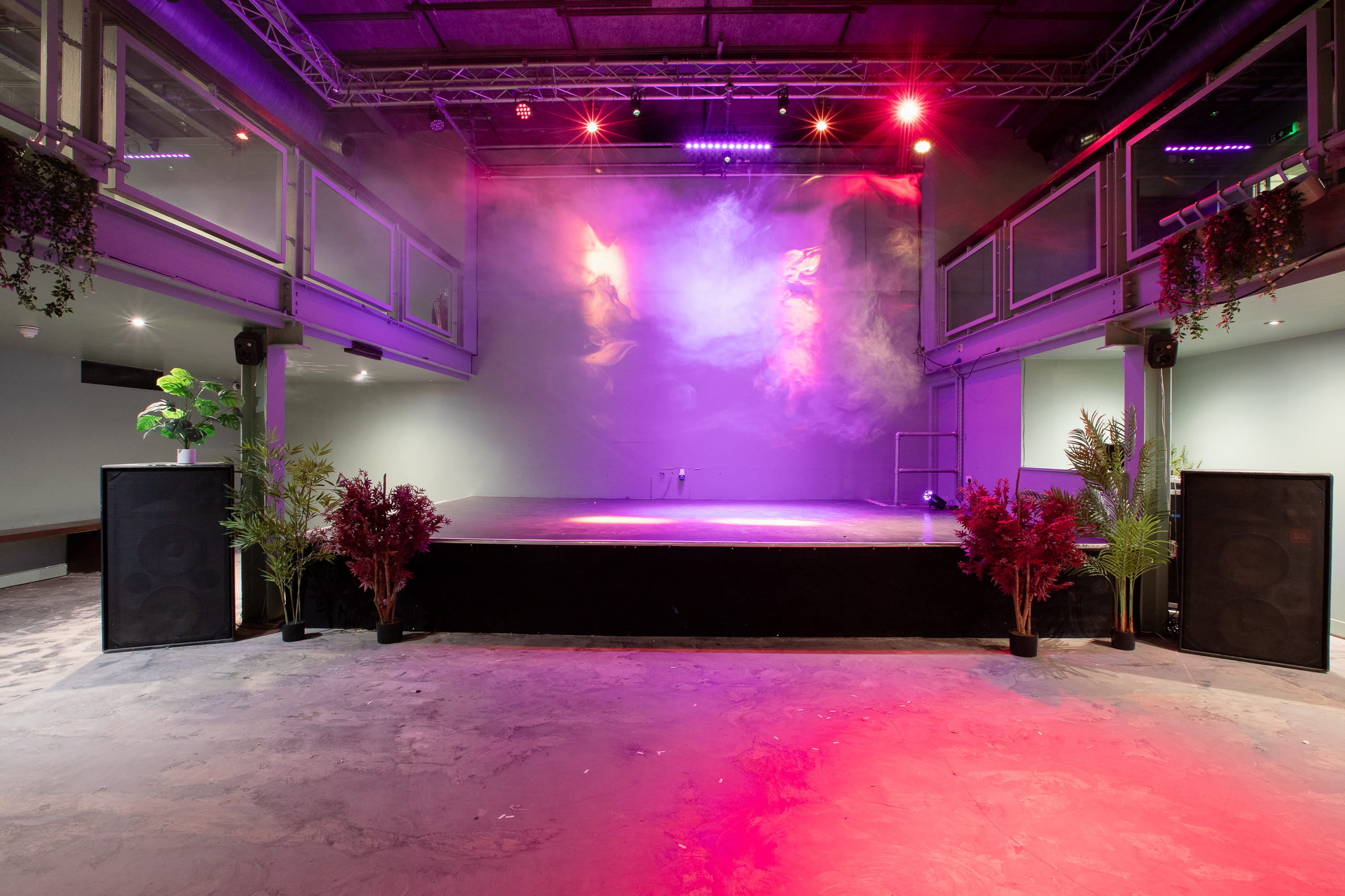 Empty stage with purple and red lighting and fog effects, surrounded by potted plants and large speakers in an indoor venue.