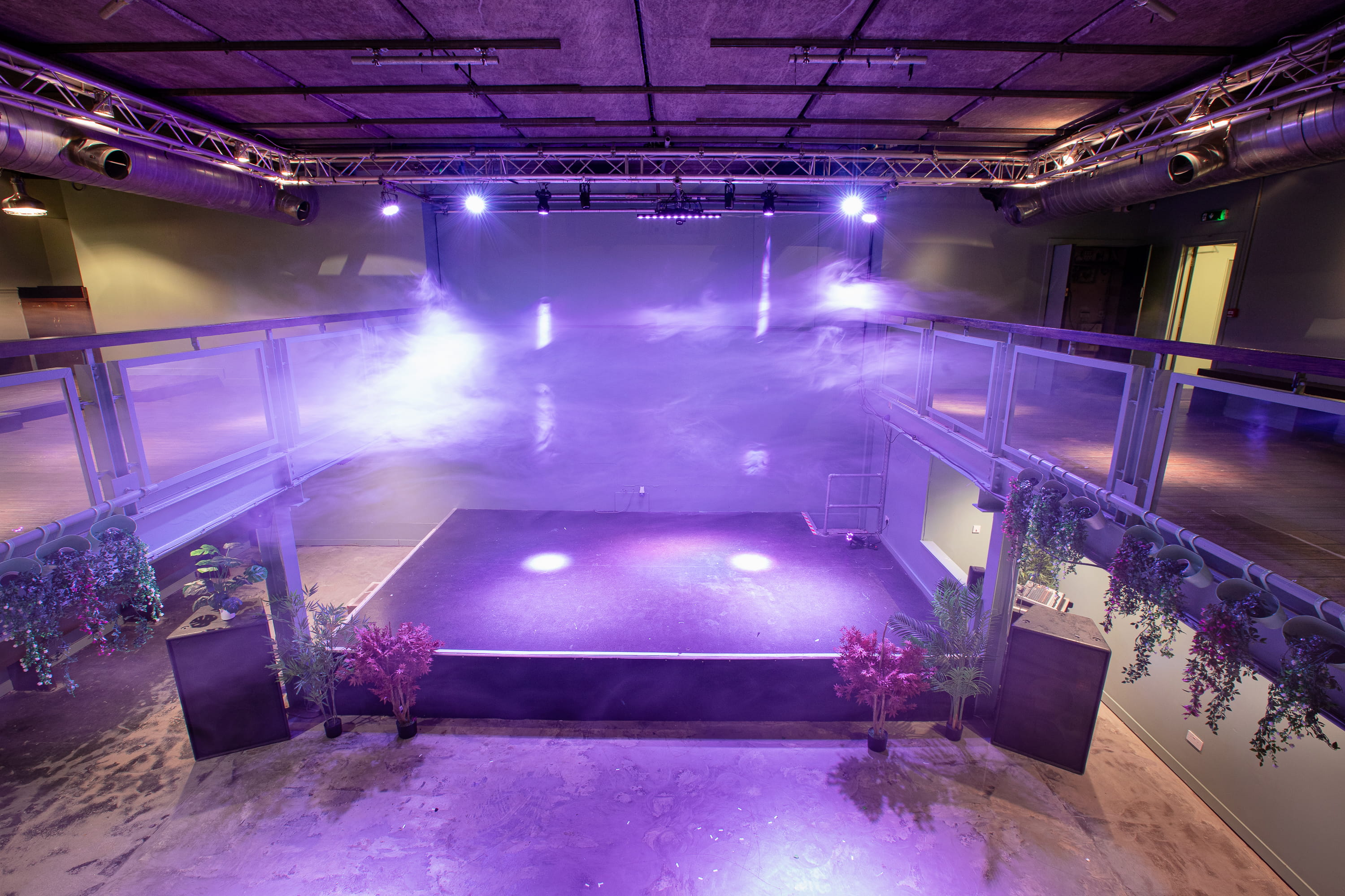 Empty indoor stage with purple lighting and fog effects, surrounded by plants and railings.
