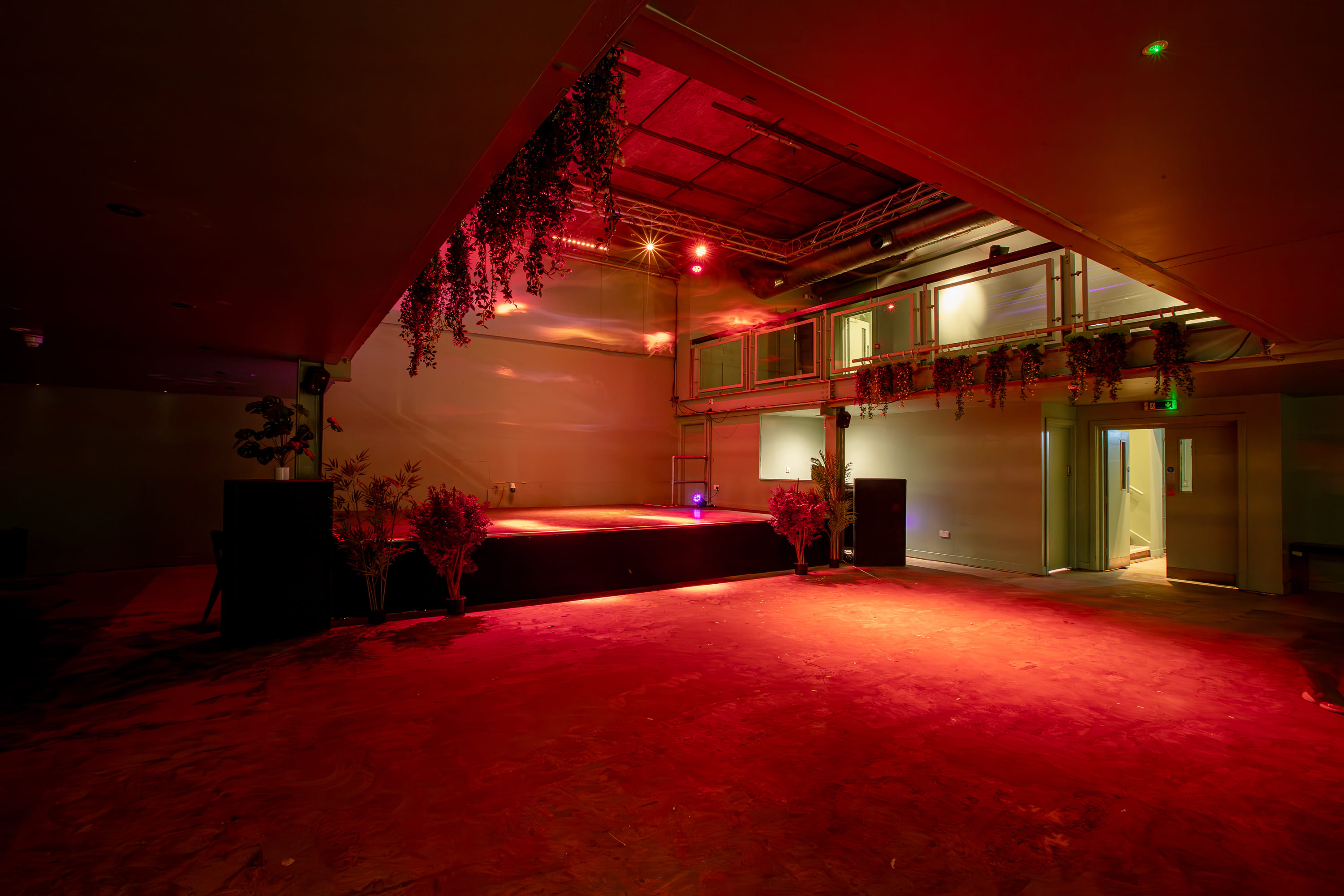 Empty indoor stage area with red lighting, hanging plants, and a balcony above.