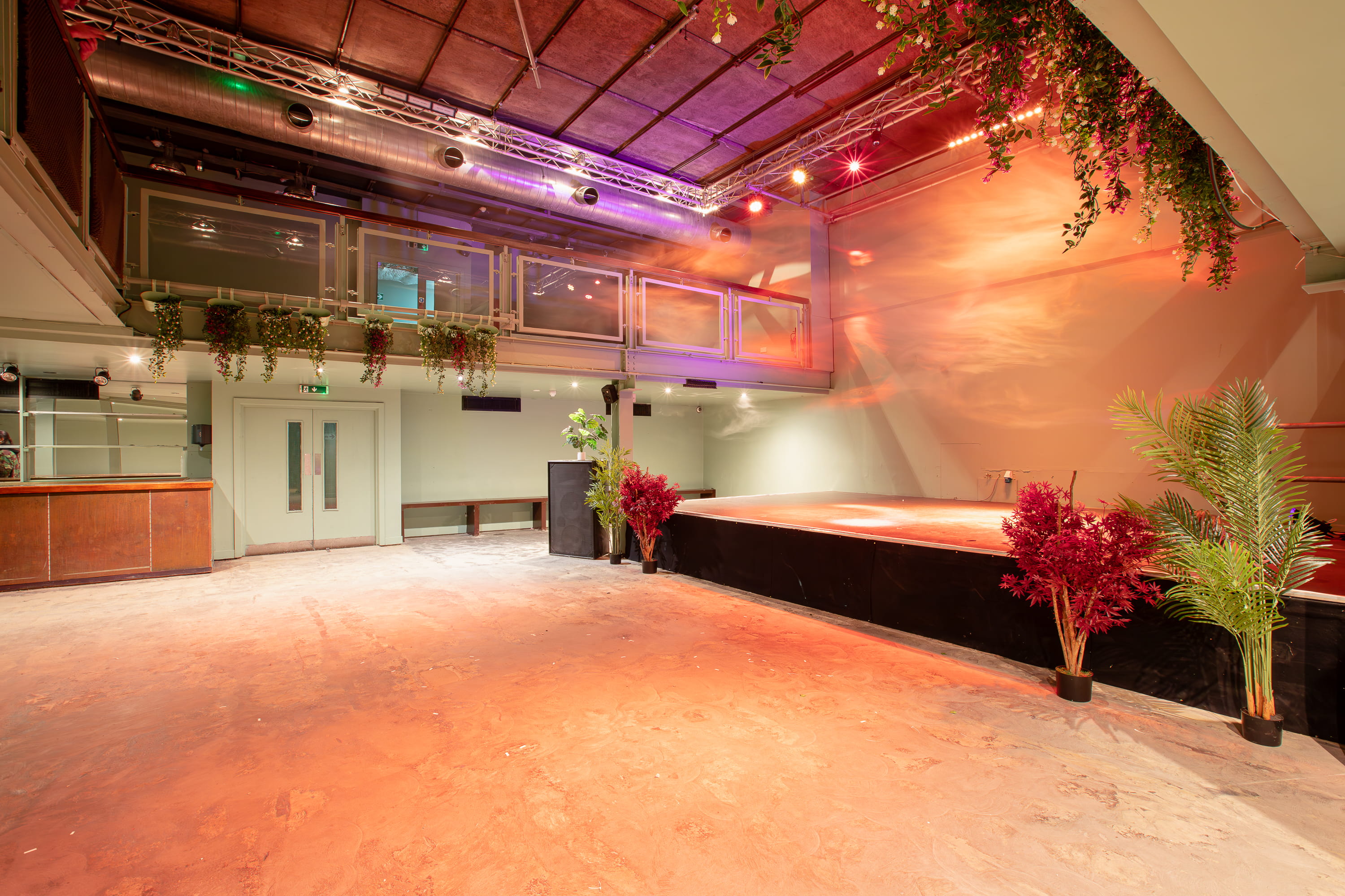 Empty indoor event space with a stage, hanging plants, and warm overhead lighting.