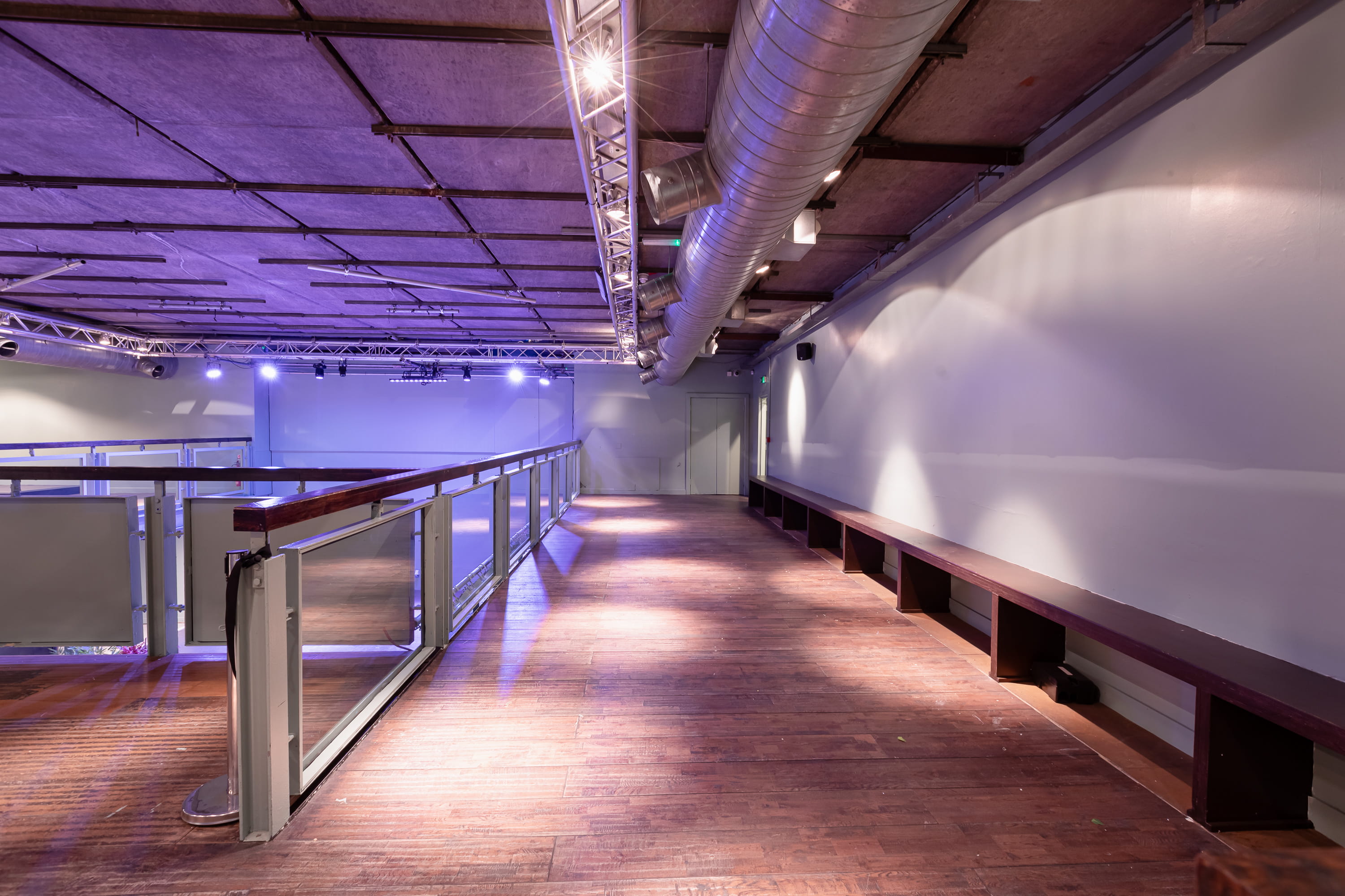 Empty indoor balcony area with wooden floor, long built-in wooden bench, glass railing, and ceiling lights.