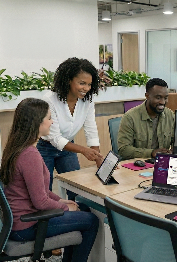 Three coworkers collaborating in a modern office, with one woman standing and pointing at a tablet while the other two sit at a desk with laptops.