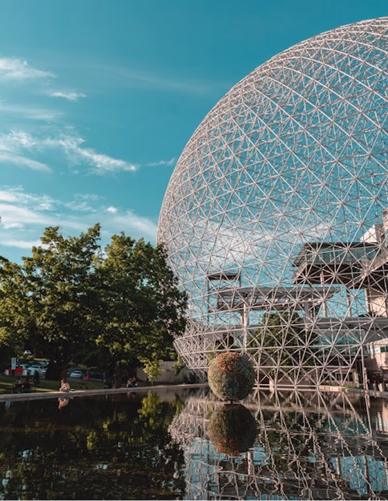 The Montreal Biosphere's geodesic dome reflecting in a still pool of water surrounded by lush green trees.