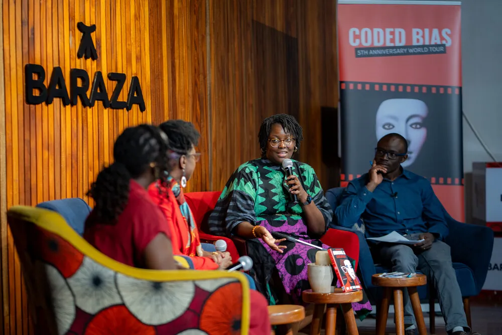 Woman in green/black patterned top speaking into microphone while Dr. Joy in red blazer and man in blue denim listen