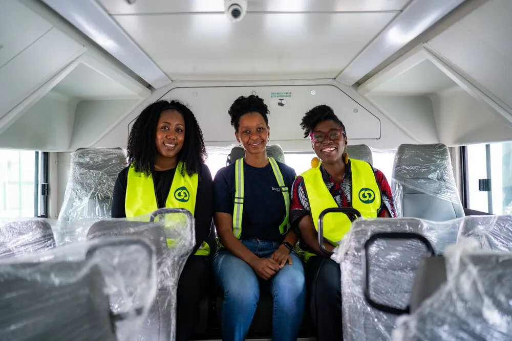 Three women smiling inside a new BasiGo electric bus with plastic-wrapped seats, two wearing yellow BasiGo safety vests