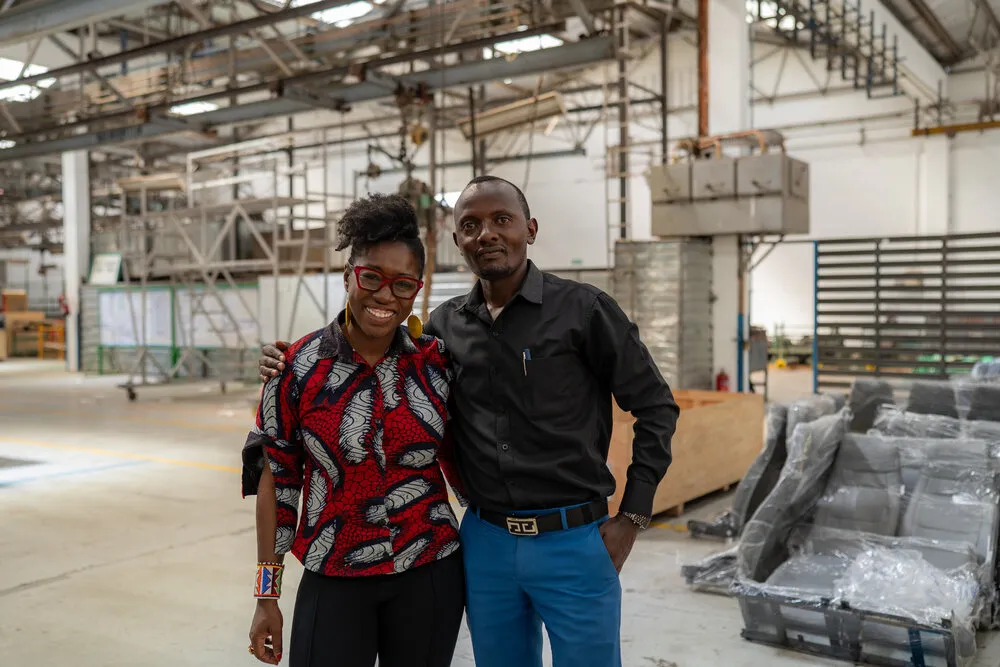 Dr. Joy in a red and black African print top smiling with a man in a black shirt in the BasiGo factory, bus seats visible